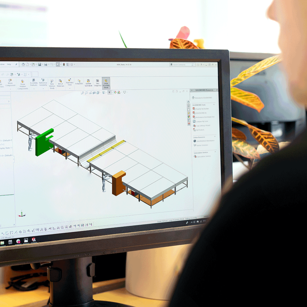 A computer monitor displays a 3D engineering model in CAD software. The workspace shows structural components and assemblies, while a person sits at the desk near the screen with plants in the background.